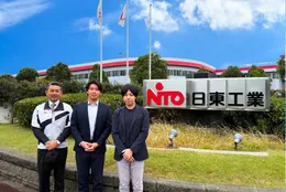 日東工業の社名看板の前に立つ日東工業とAPTの担当者